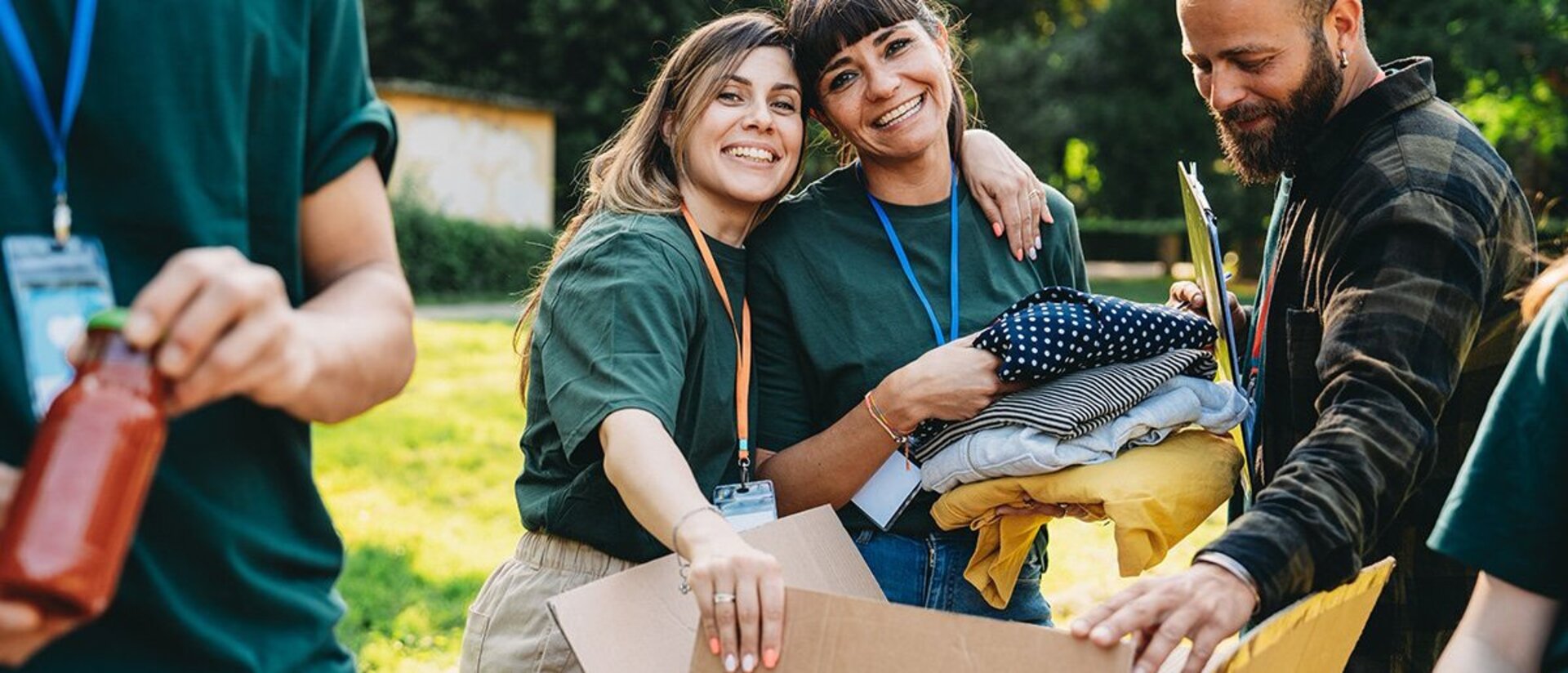 Mehrere Personen in den Zwanzigern tragen Namenschilder an Bändern um den Hals Sie haben einen geöffneten Karton vor sich. Die eine Frau in der Mitte legt der Frau neben sich den Arm um die Schulter. Diese hält mehrere Wäschestücke im Arm. Der Mann rechts trägt die Haar zusammengebunden zu einem Zopf und hält den Karton auf. 